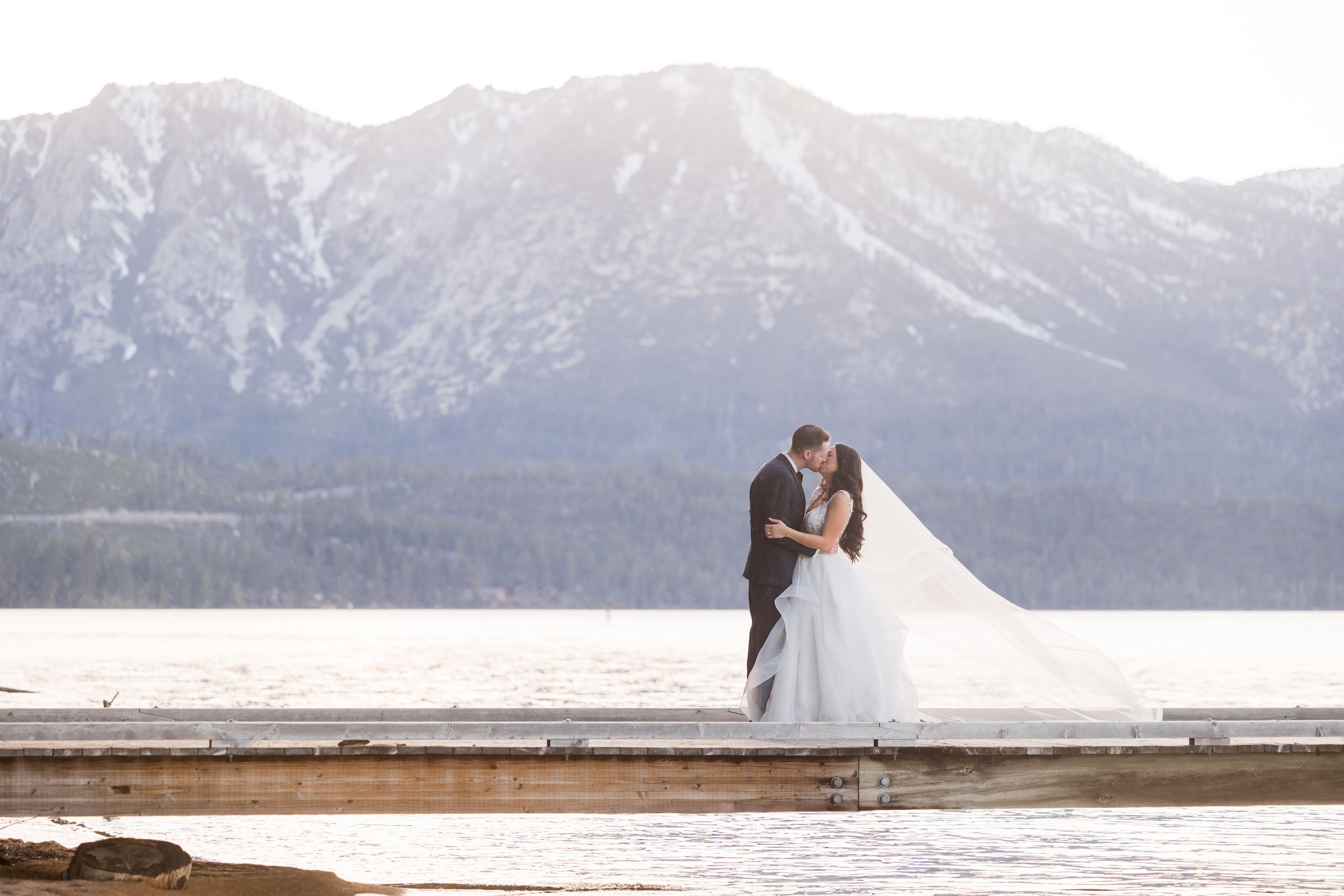 Bride and groom share a kiss on a dock with snow-capped mountains and a serene lake in the background.