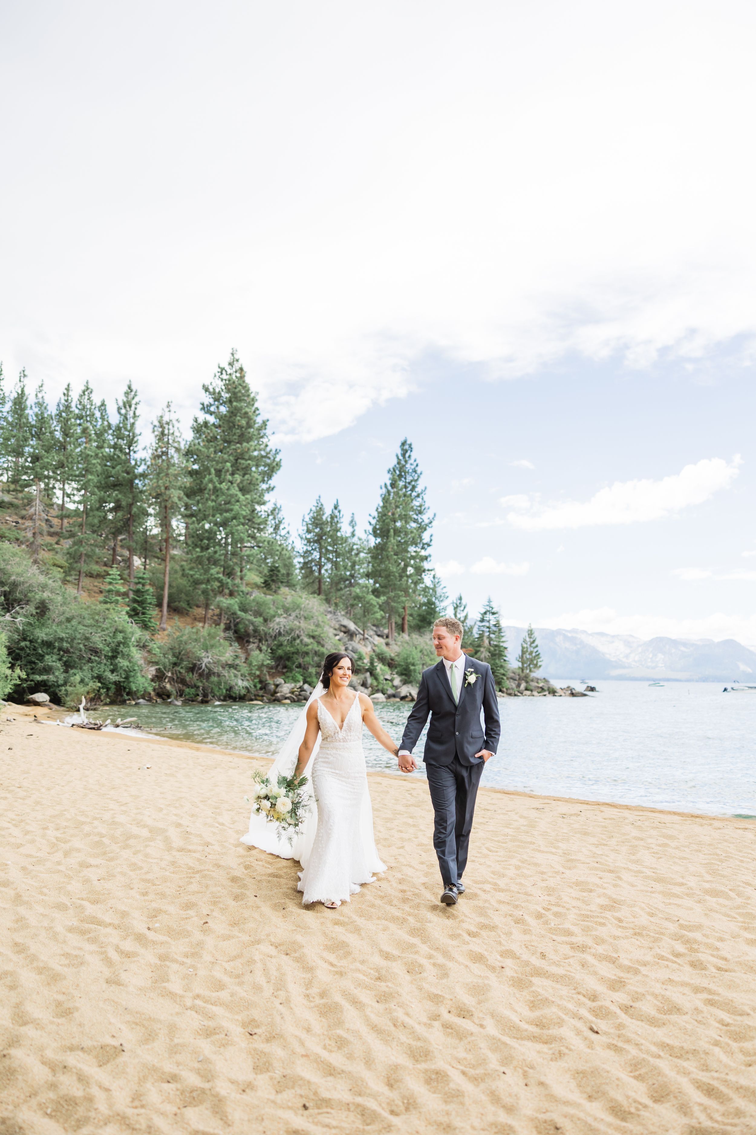 Bride and groom walk hand-in-hand on sandy Lake Tahoe beach, surrounded by pine trees and mountain views.