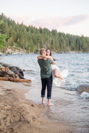 Couple embracing on a sandy lakeside beach at sunset, surrounded by pine trees and gentle waves.