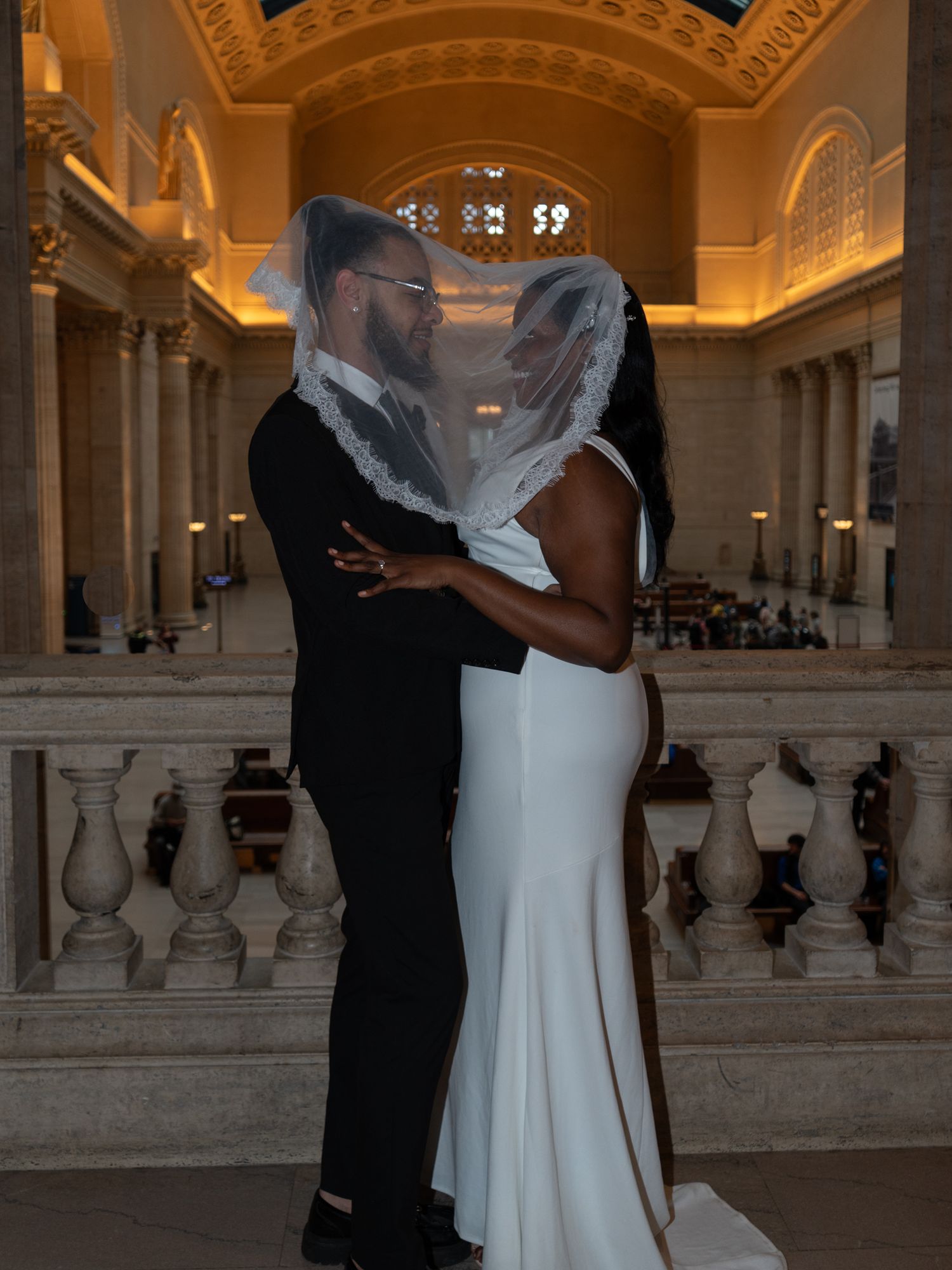 A couple shares a romantic moment in an elegant wedding venue, the bride in a white gown with veil, groom in a black tuxedo.