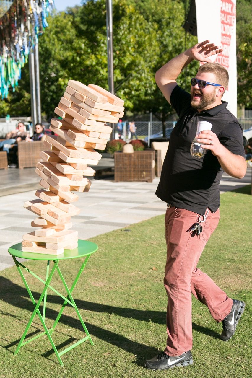 Man reacts with surprise as giant Jenga tower falls during outdoor lawn game event.