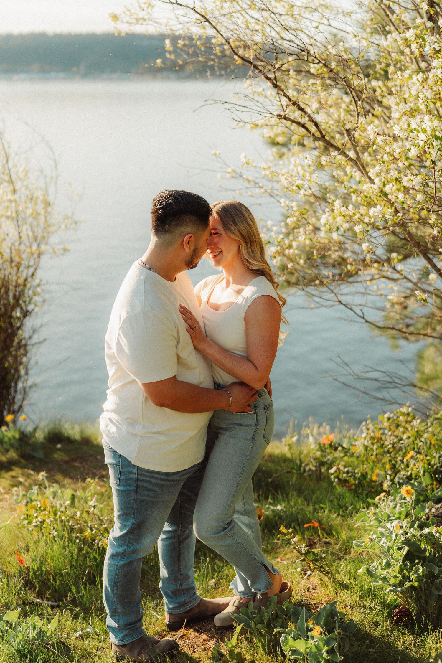Couple embracing near a sunlit lake surrounded by blooming spring trees and wildflowers.