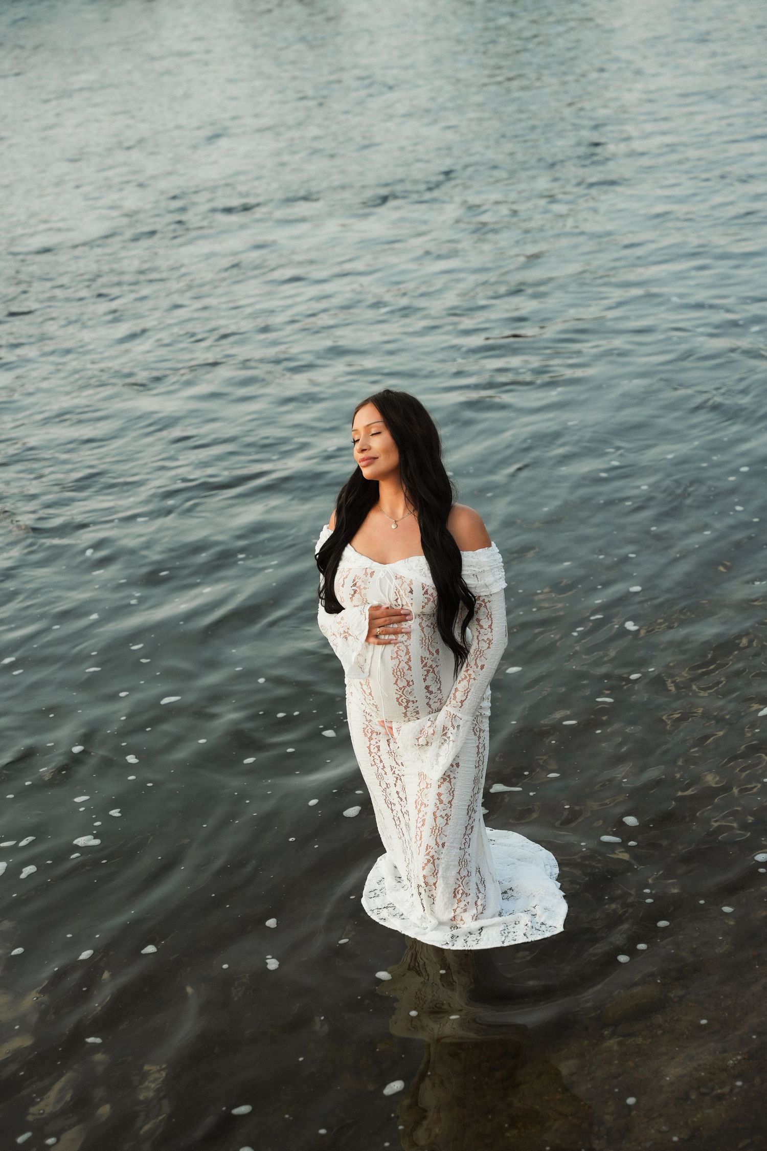 Pregnant woman in white lace off-shoulder gown standing in calm water, long dark hair, serene expression.