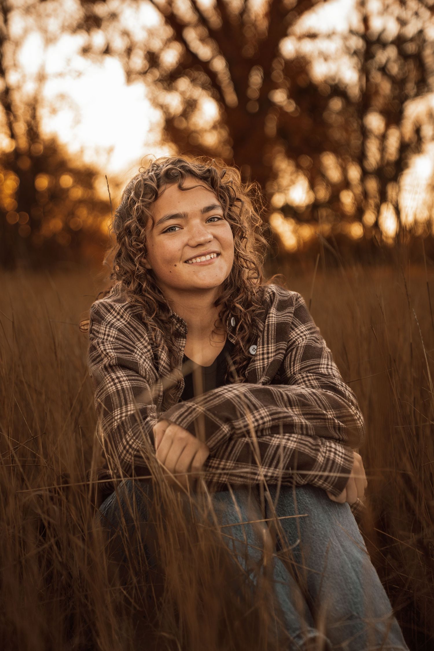 Smiling young woman with curly hair sitting in golden field grass wearing plaid shirt at sunset.