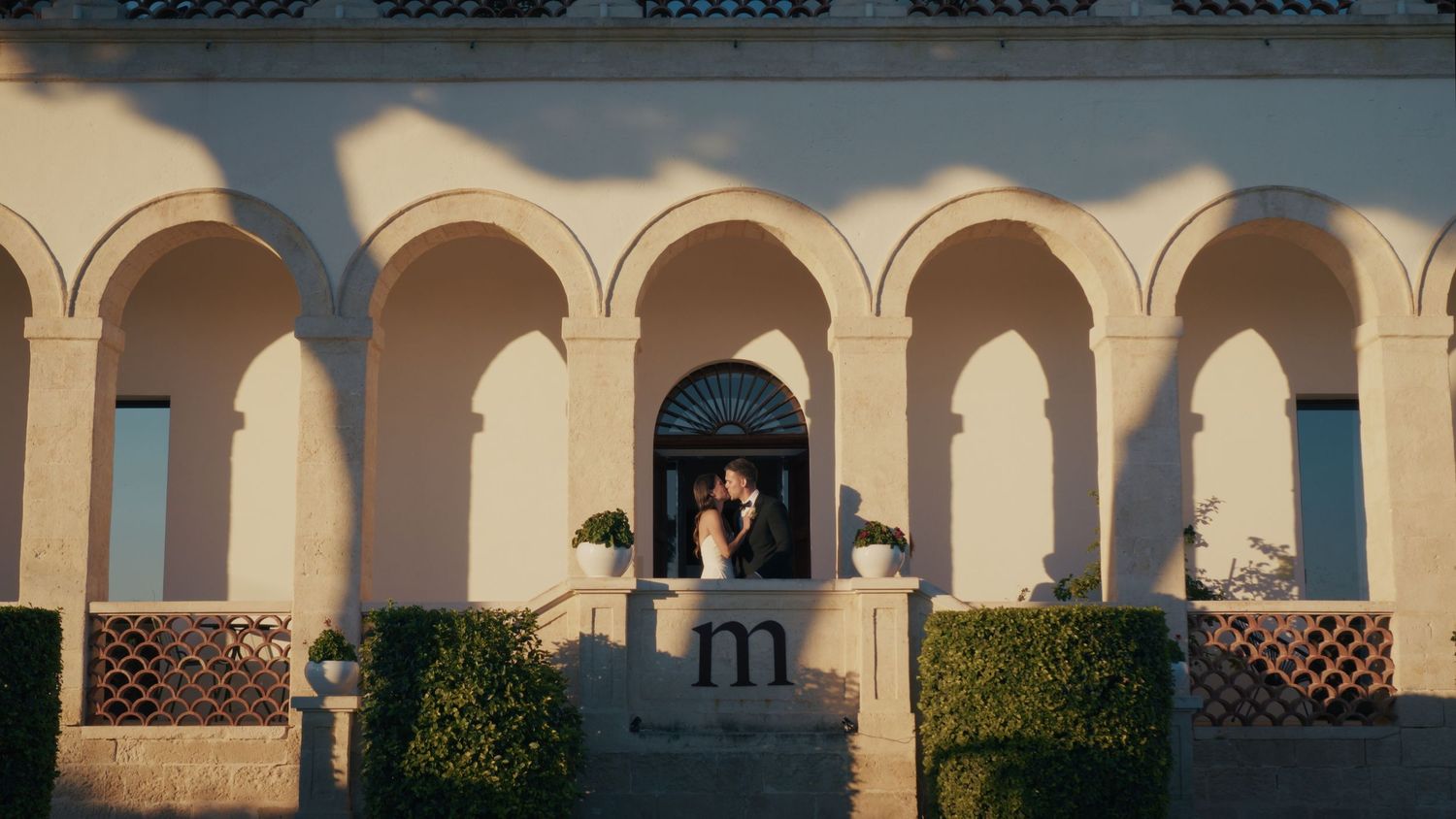 Elegant wedding couple standing under arched colonnade of a classic Mediterranean villa with lush greenery.