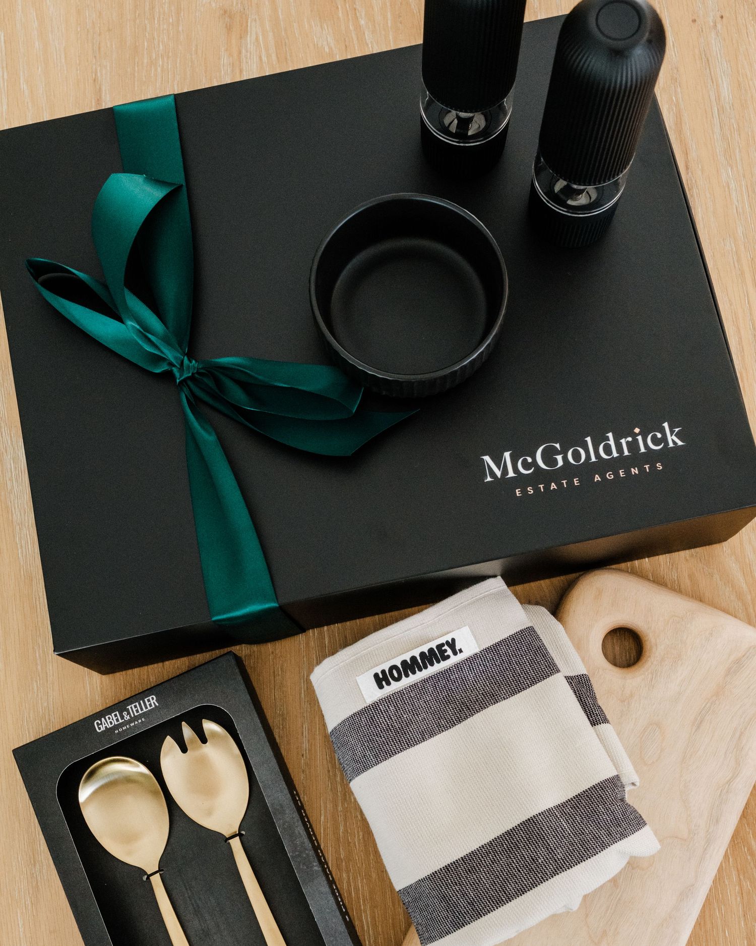 Black McGoldrick Estate Agents gift box with gold salad servers, striped tea towel, pepper grinders, and teal ribbon bow.