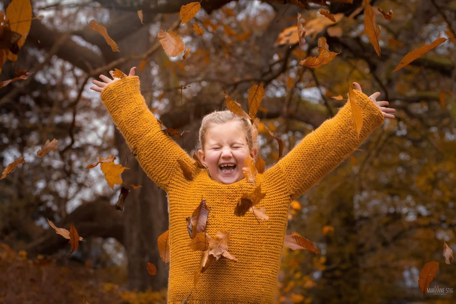 A joyful girl in a mustard sweater laughs with arms raised as autumn leaves fall around her in a park.