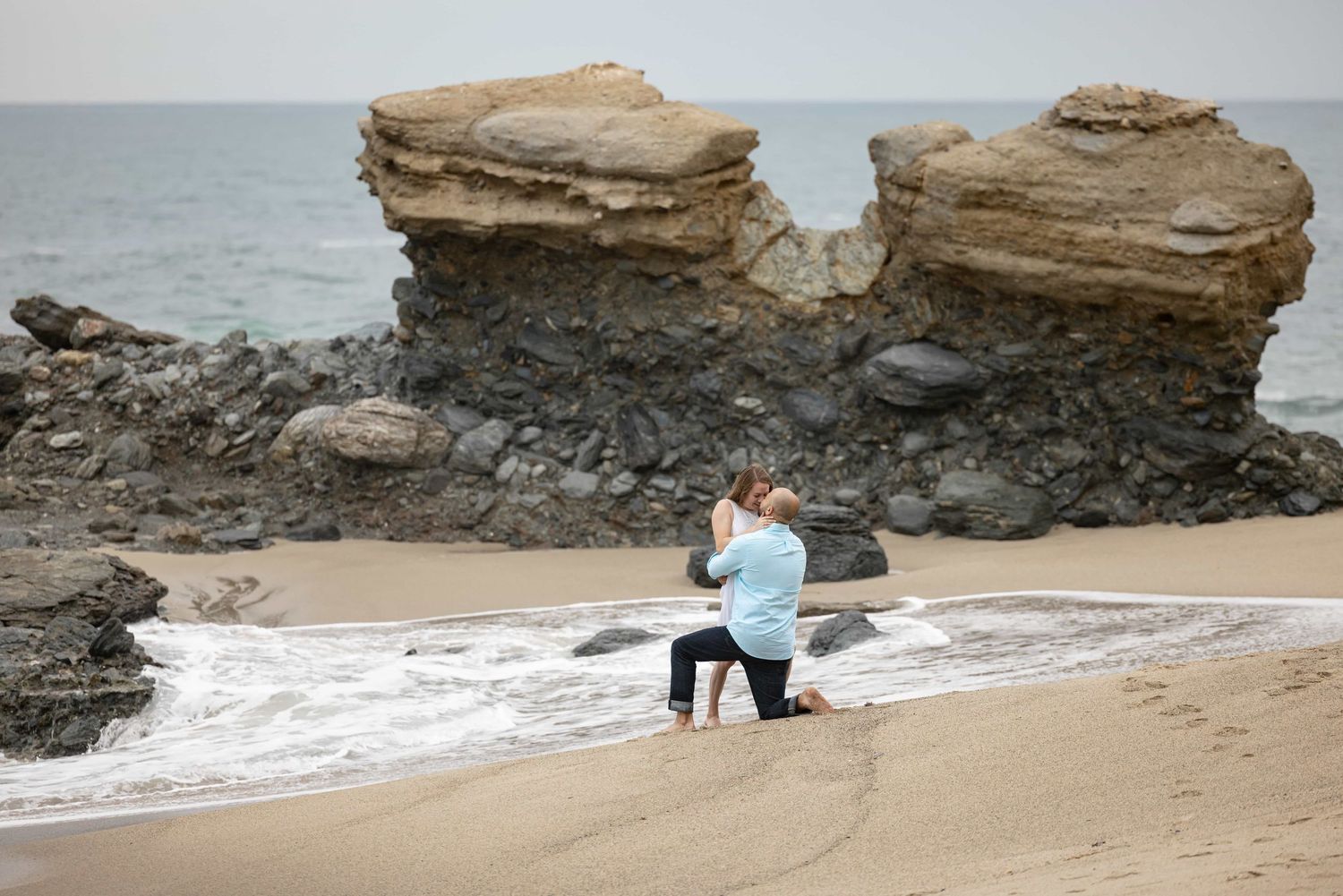 Man proposing on knees on sandy beach with rocky cliffs.