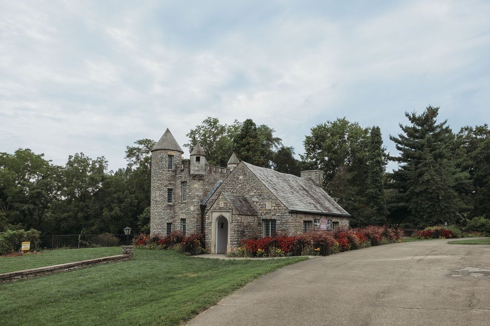 Historic stone chapel with turret surrounded by lush green trees and colorful flower beds on a cloudy day.