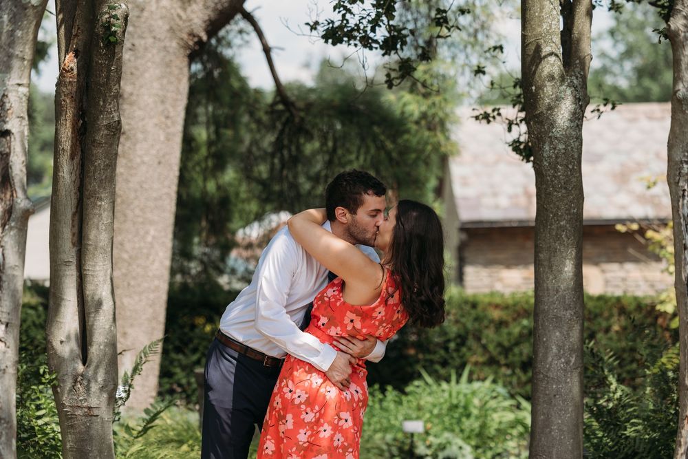 Couple sharing a romantic kiss among trees in a lush garden, woman wearing a red floral dress.