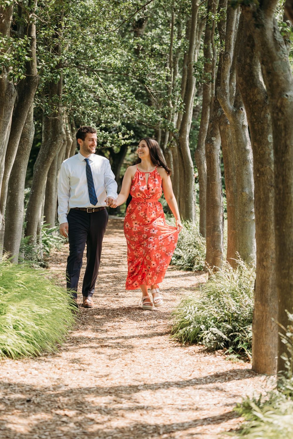 Couple holding hands walking along a tree-lined path, woman in red floral maxi dress, man in white shirt and navy pants.