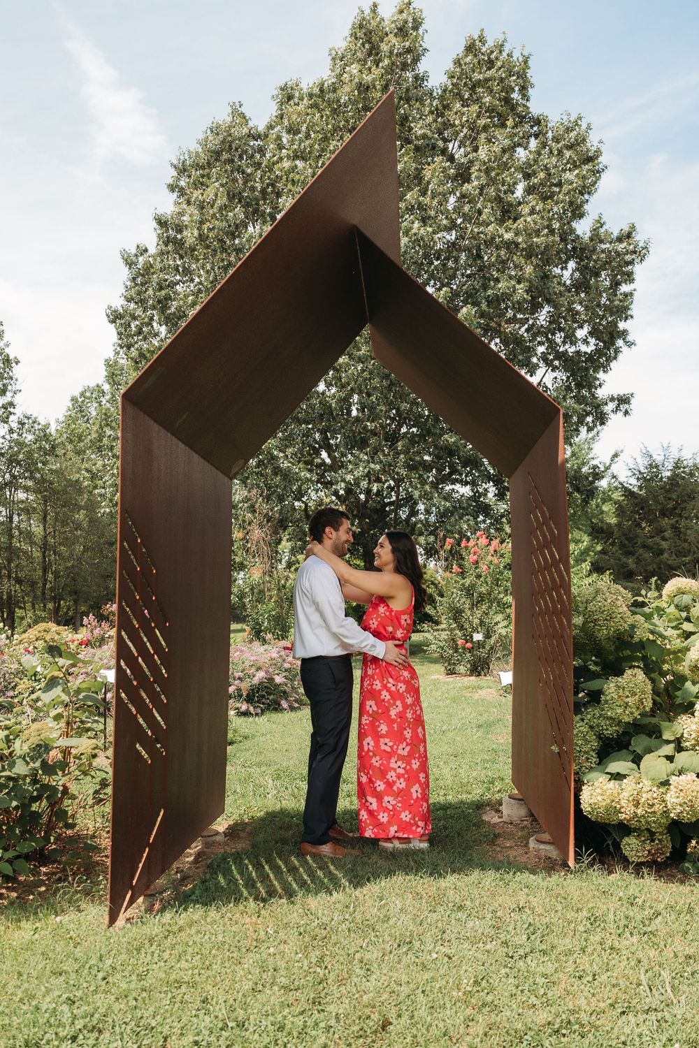 Couple embracing under a rustic metal arch sculpture in a lush garden with blooming flowers.