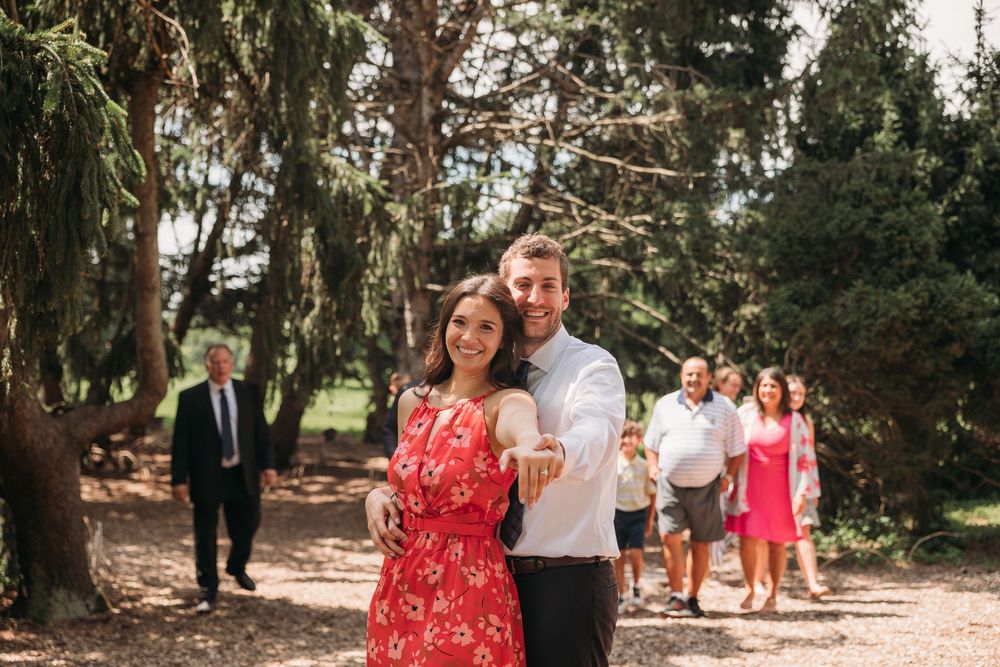 Smiling couple in red floral dress and black shirt posing outdoors in a park with guests in the background.
