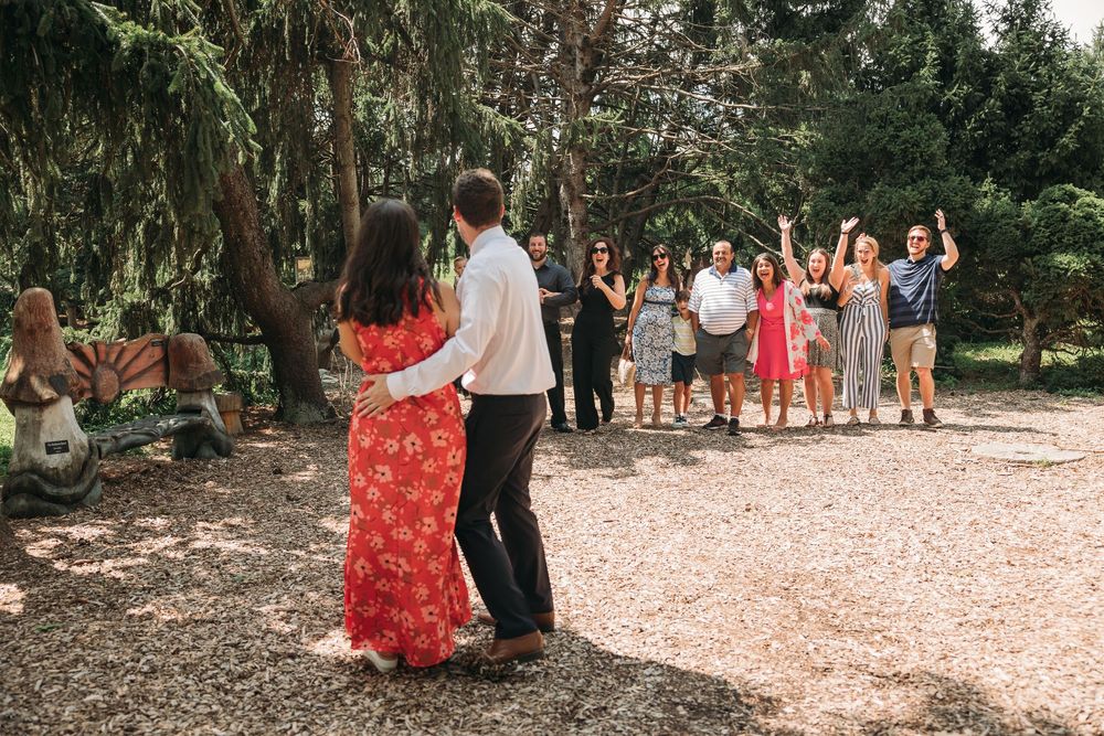 Couple dancing outdoors while smiling family and friends celebrate in background under lush green trees.