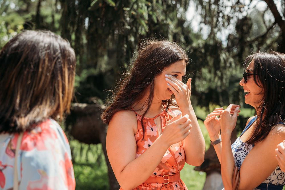 Emotional woman in floral dress covering her mouth in surprise, surrounded by friends outdoors.