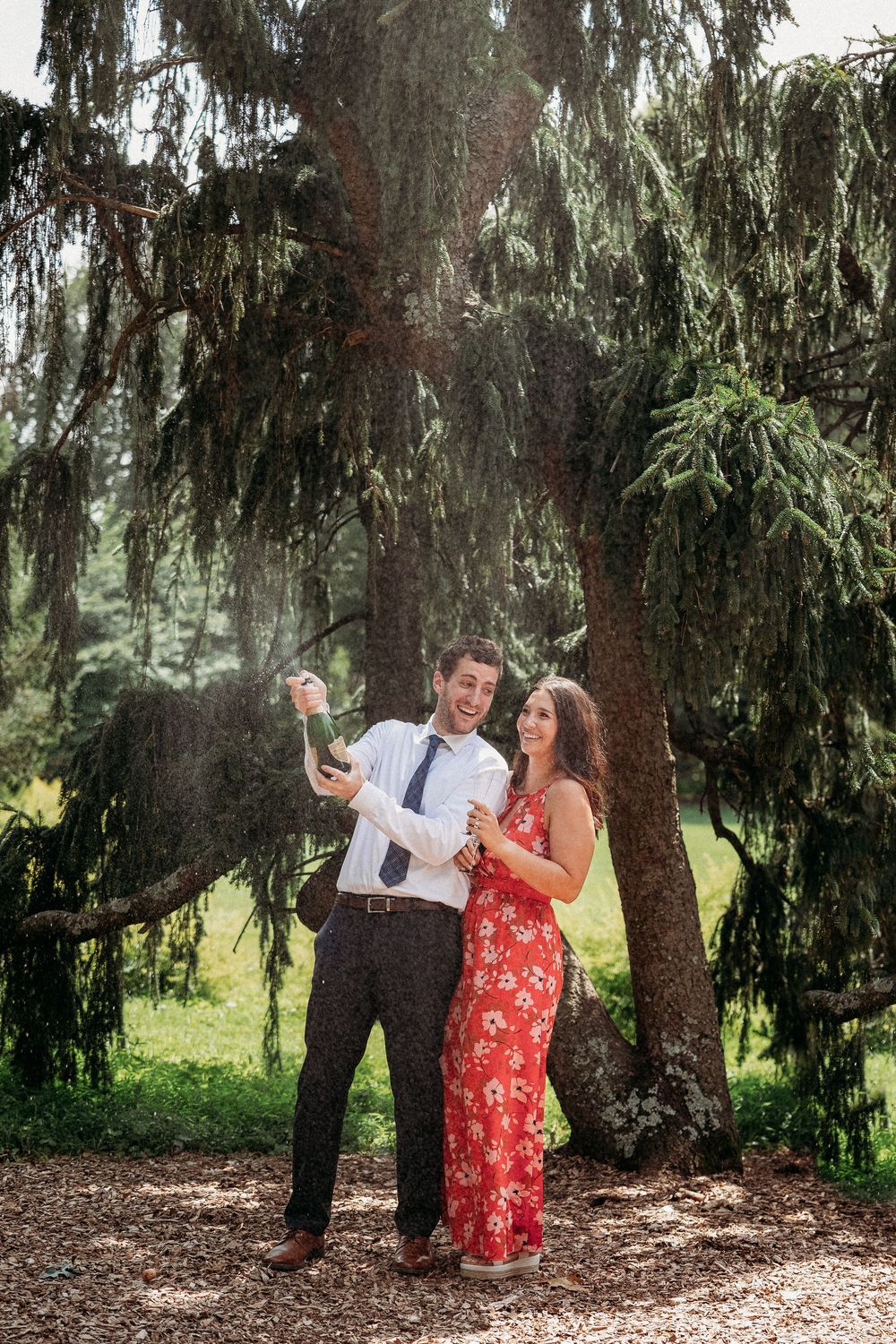 Couple celebrating outdoors under pine trees, man popping champagne bottle, woman in red floral dress smiling.