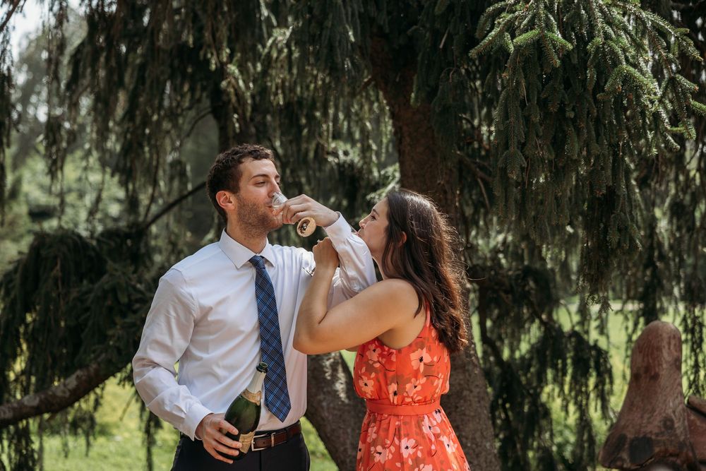 Couple sharing a champagne bottle toast outdoors under a weeping willow tree, woman in red floral dress.