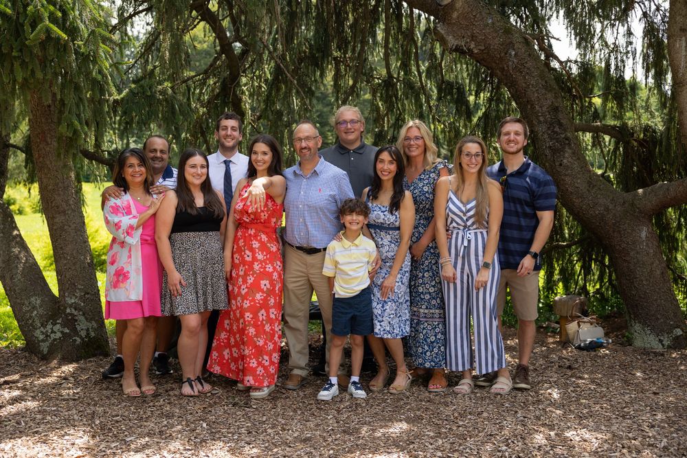 Large family group posing together outdoors under weeping willow trees on a sunny summer day.