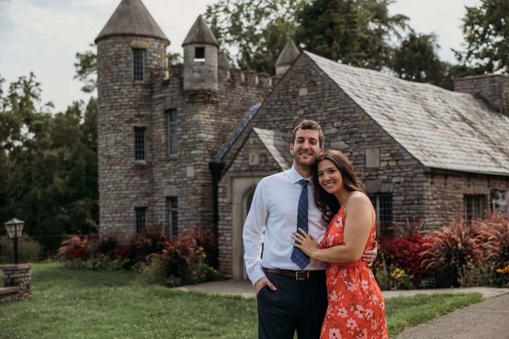 Couple smiling in front of a stone castle venue surrounded by lush greenery, man in blue suit, woman in floral dress.