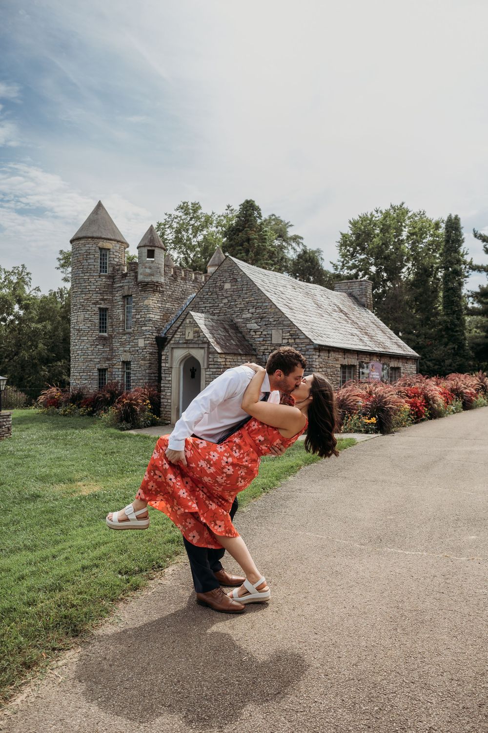 Couple sharing a romantic dip kiss in front of a charming stone castle building surrounded by colorful gardens.