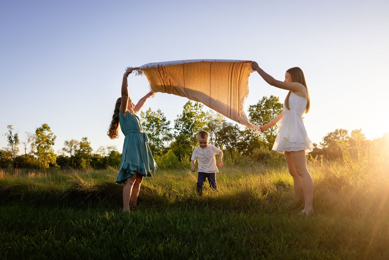 Two women hold a blanket over a toddler in a sunny green field at golden hour.