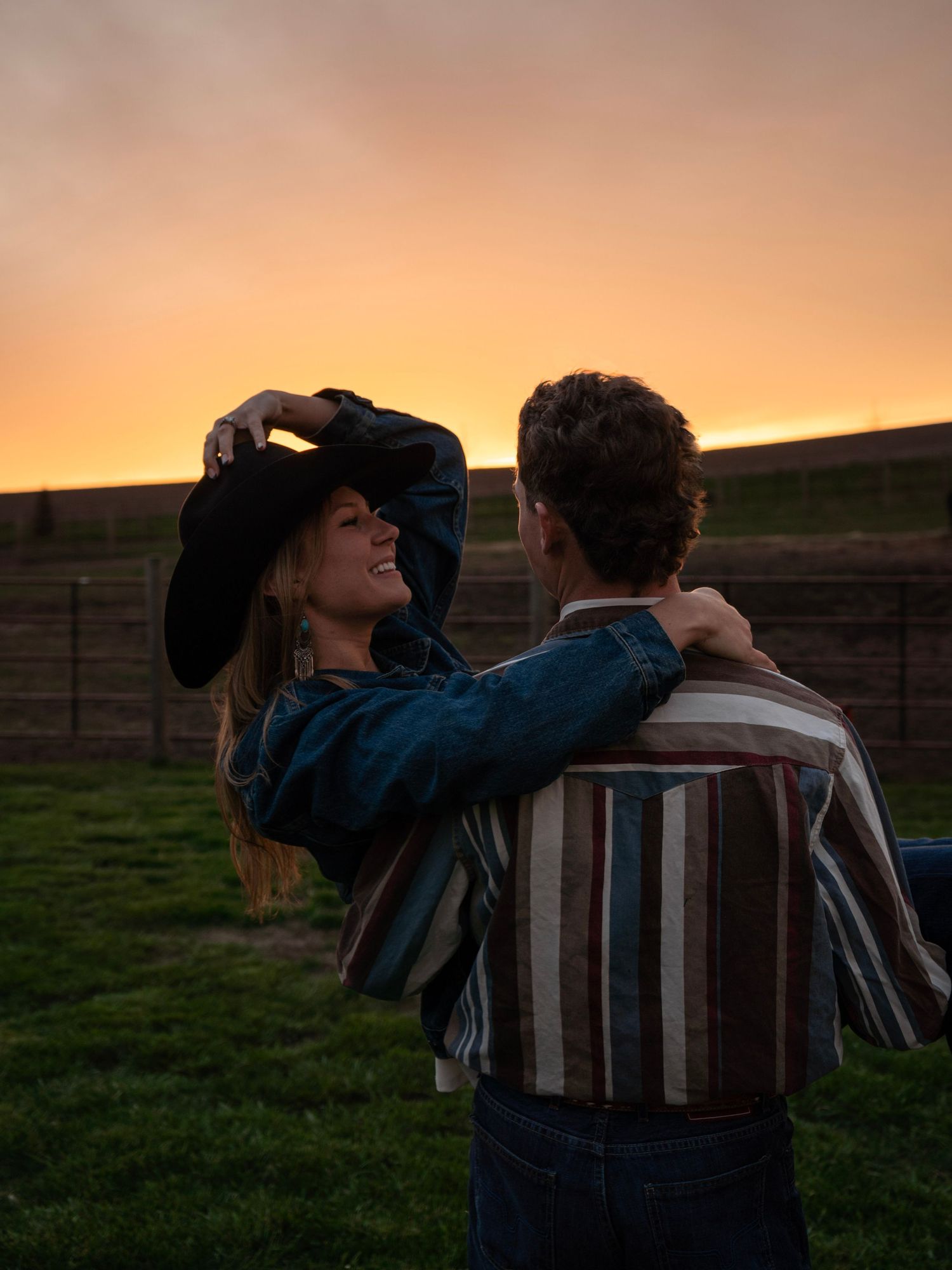 Couple dancing at sunset in a field, woman in denim jacket and cowboy hat, man in striped western shirt.