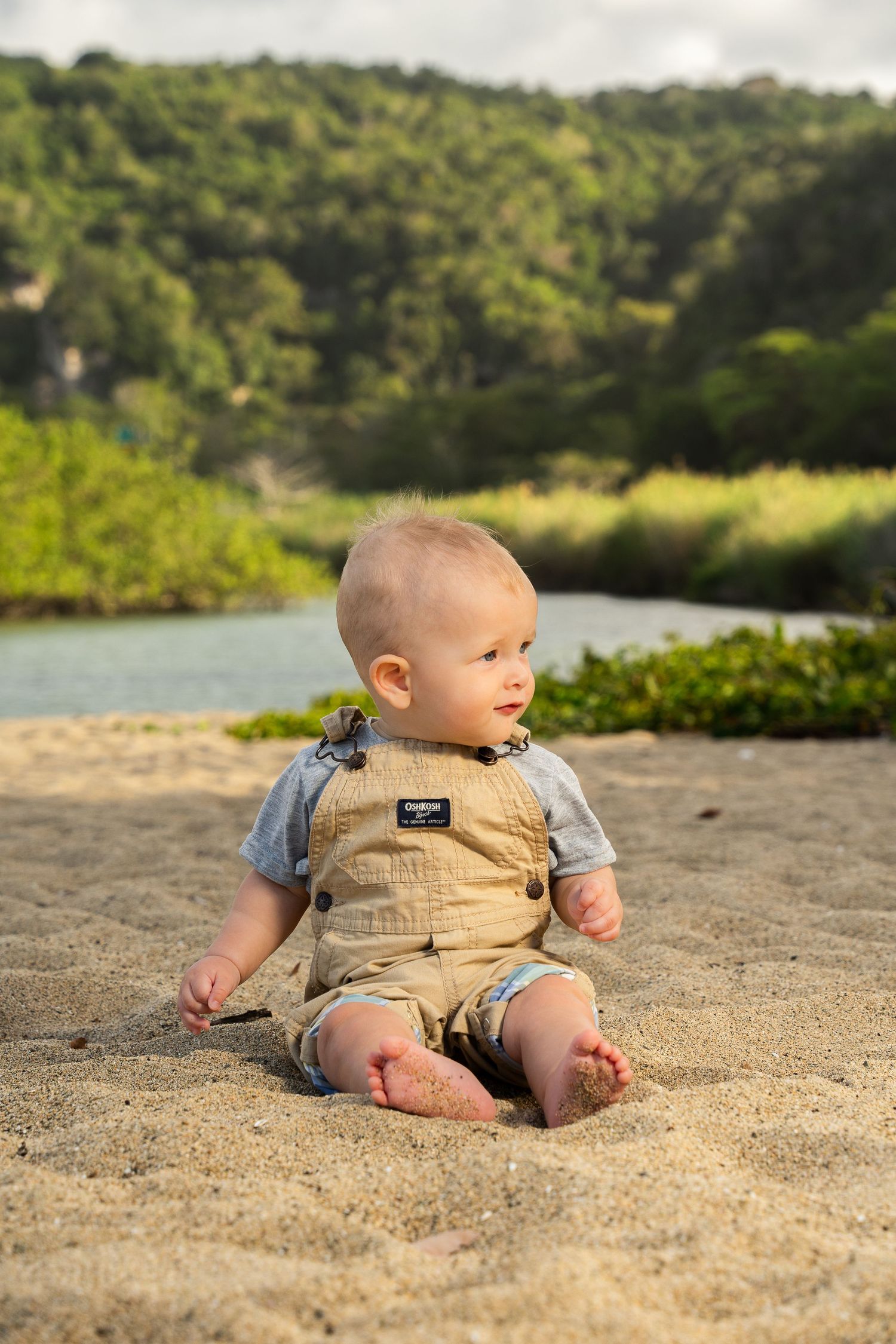 Adorable baby in tan overalls sitting on sandy beach with lush green trees and river in background.