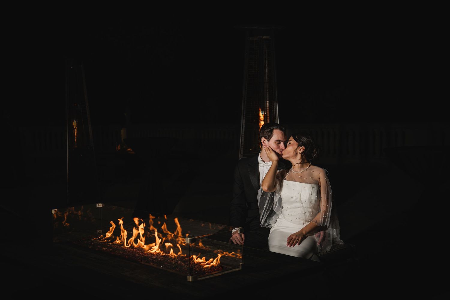 Romantic couple embracing beside glowing outdoor fire pit in dark evening wedding setting.