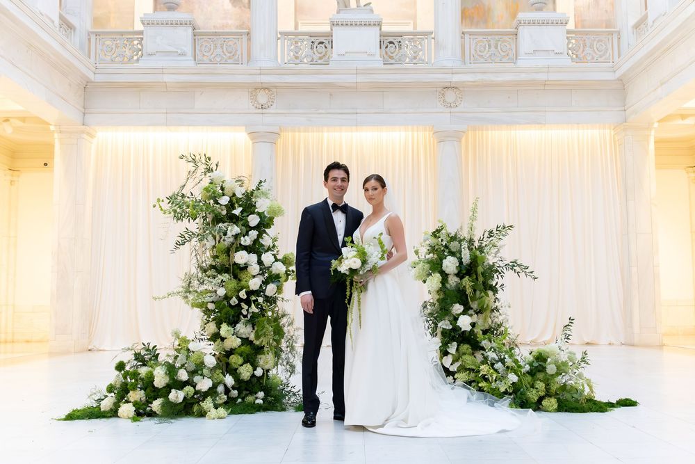 Bride and groom stand before lush white floral arrangements in an elegant white marble wedding venue.