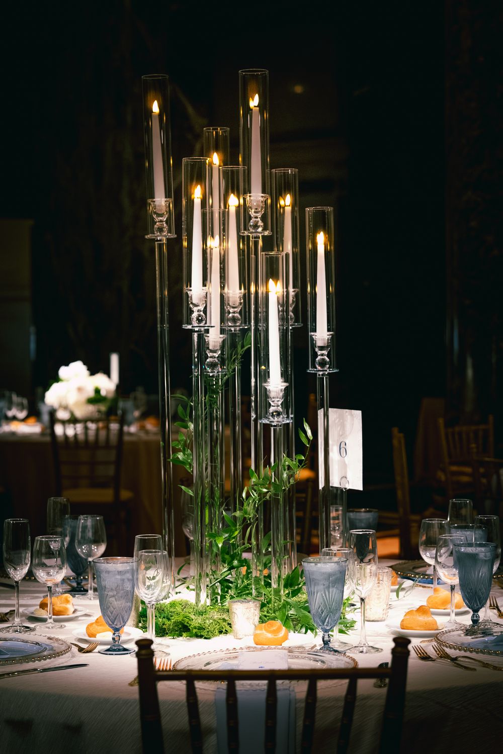 Elegant wedding table centerpiece with tall glass candelabra, lit candles, greenery, and gray stemware at table six.