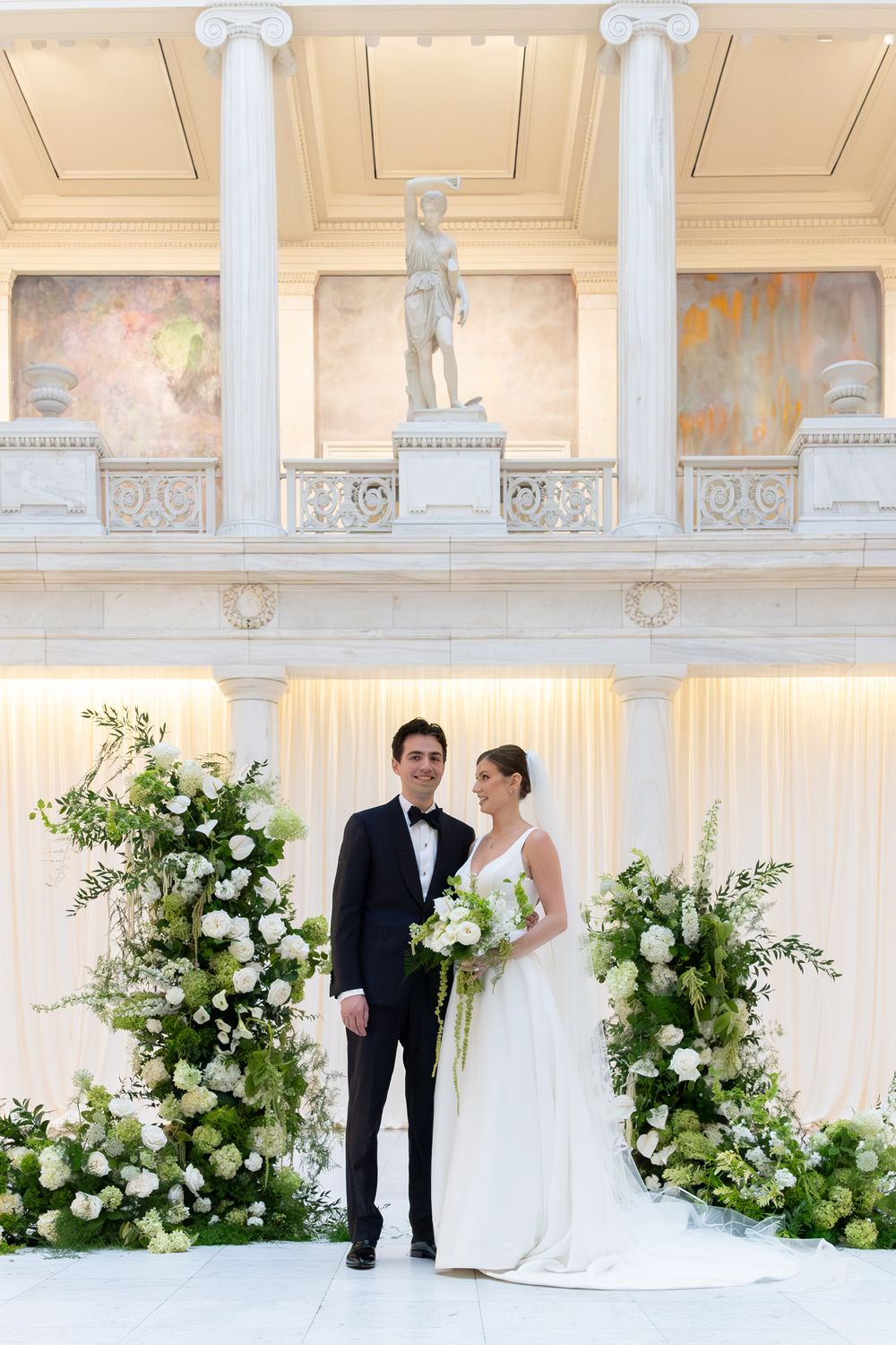 Bride and groom pose before white floral arch columns in elegant neoclassical venue with marble statue backdrop.