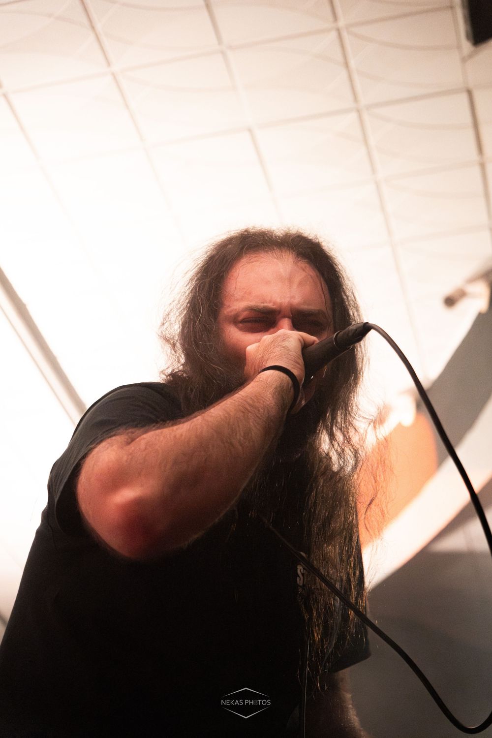 Long-haired metal singer performing intensely into microphone under bright white ceiling stage lights.