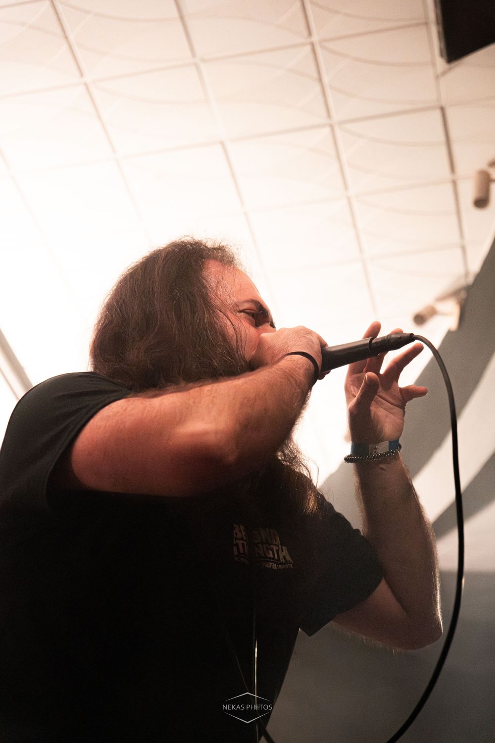 Long-haired male rock vocalist performing on stage, singing intensely into a microphone while wearing a black band t-shirt.