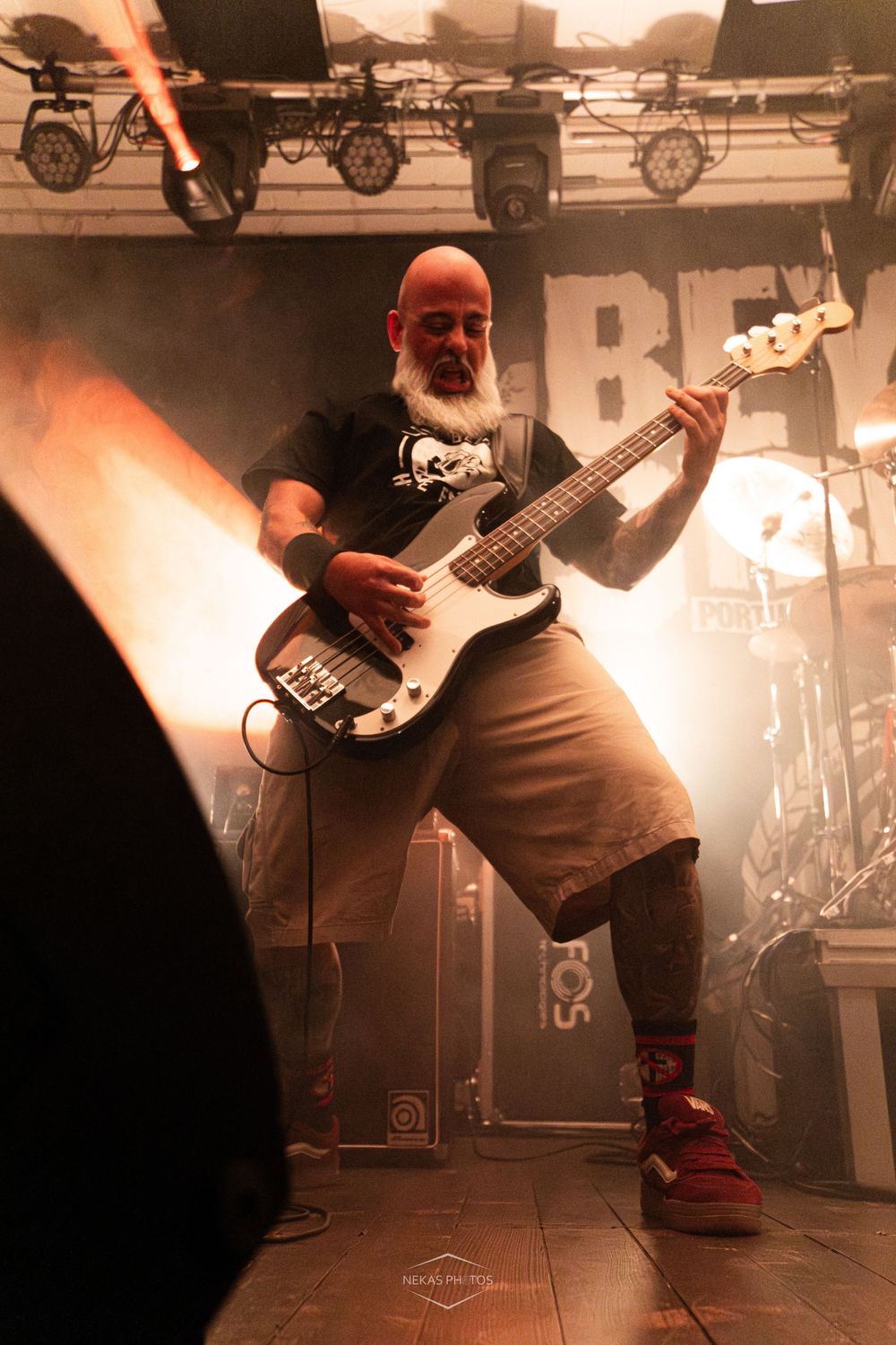 Bald bearded bassist performs on smoky stage, playing electric bass guitar in black t-shirt and khaki shorts at rock concert.