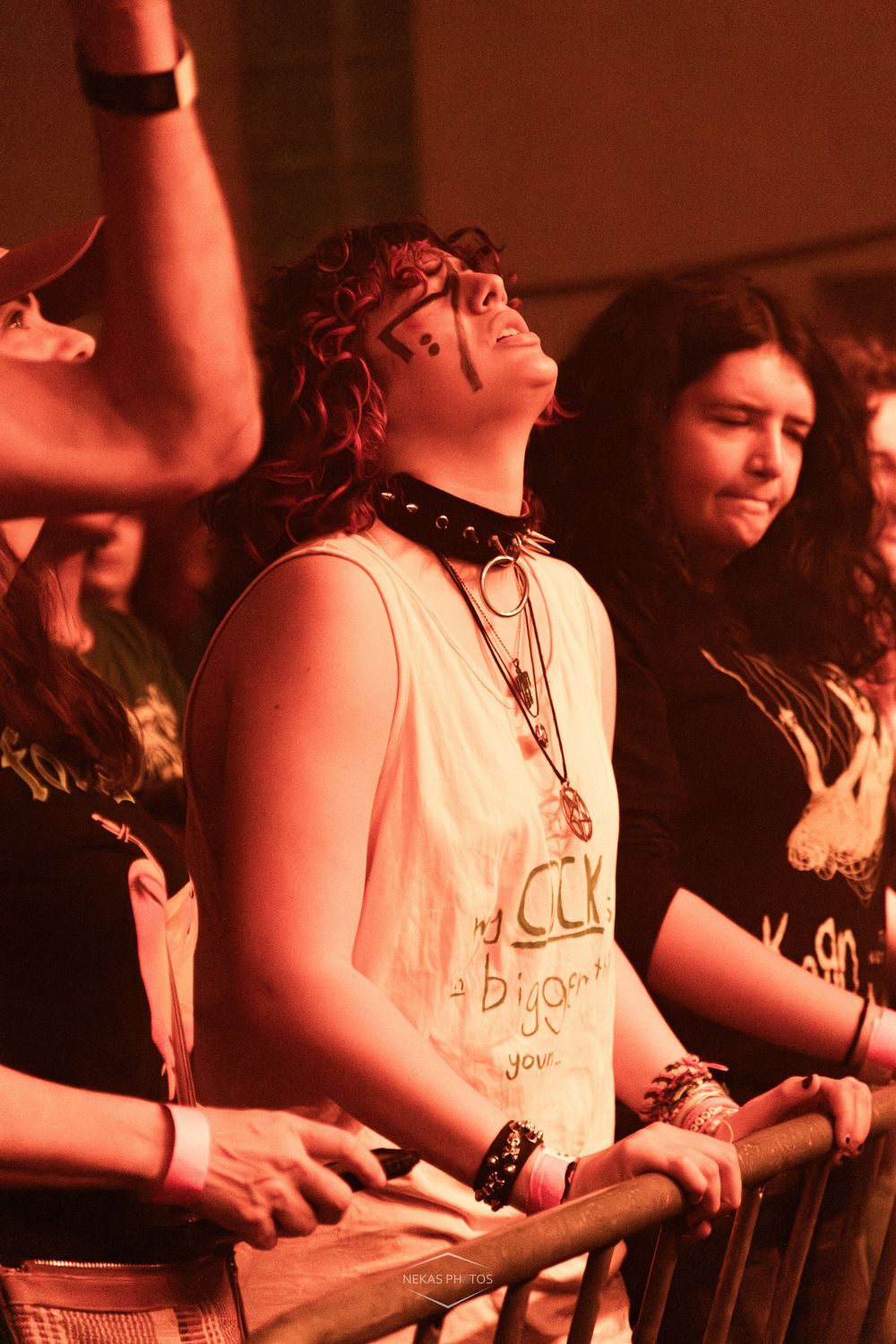 Concert crowd with fans leaning on barrier under red stage lighting, woman in white tank top and choker looking up.
