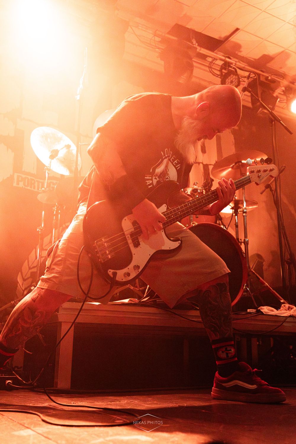 Bald guitarist performs intensely on stage bathed in warm orange light, leaning over electric bass guitar at live rock concert.