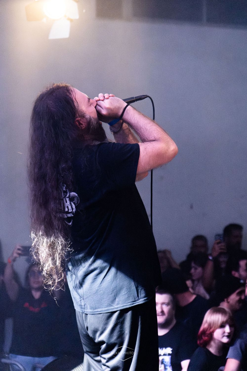 Long-haired metal vocalist performs passionately on stage, holding microphone close, with crowd visible in background.