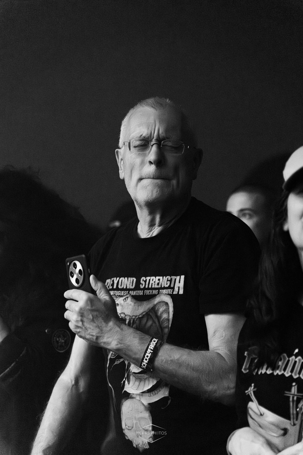 Black and white photo of an older man wearing glasses and a Beyond Strength t-shirt holding a phone at a concert.