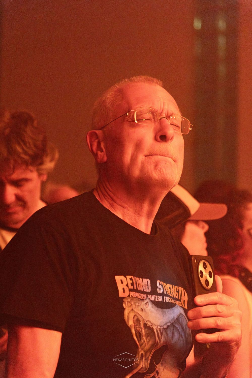 Older man with glasses wearing a Beyond Strength black t-shirt holds a phone at a red-lit event.