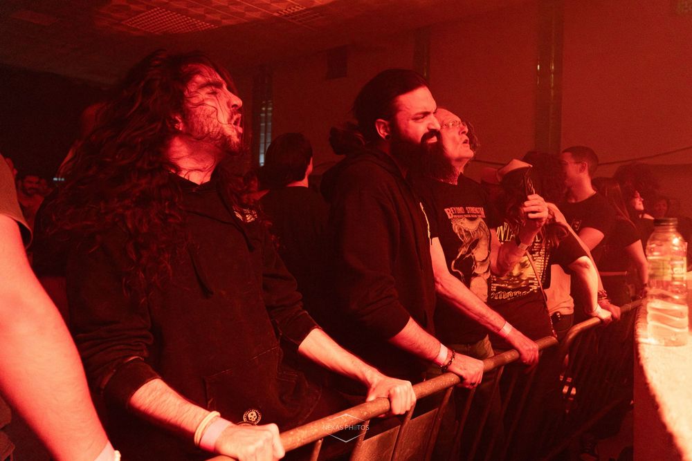 Crowd of metal fans bathed in red light enjoying a live concert, standing at the barrier with intense expressions.