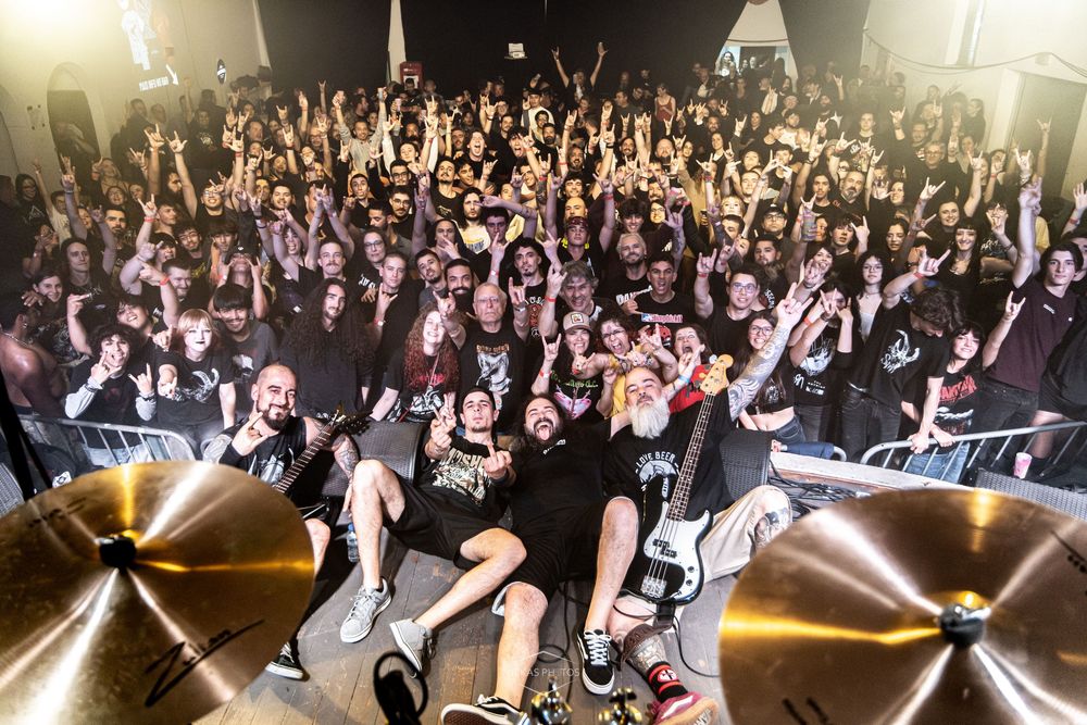 A metal band poses on stage with a massive crowd of fans cheering behind them at a live concert.