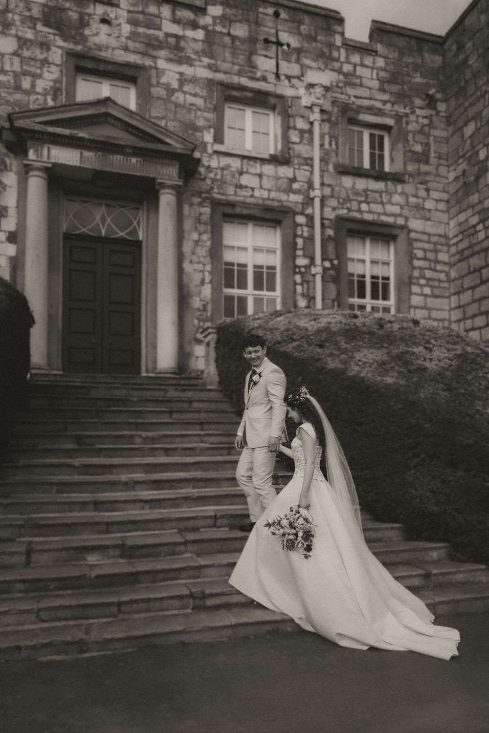 Black and white photo of bride and groom posing on stone steps outside a historic church building.