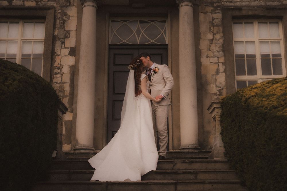 A bride and groom share a romantic moment on stone steps outside an elegant manor, her white veil flowing gracefully.