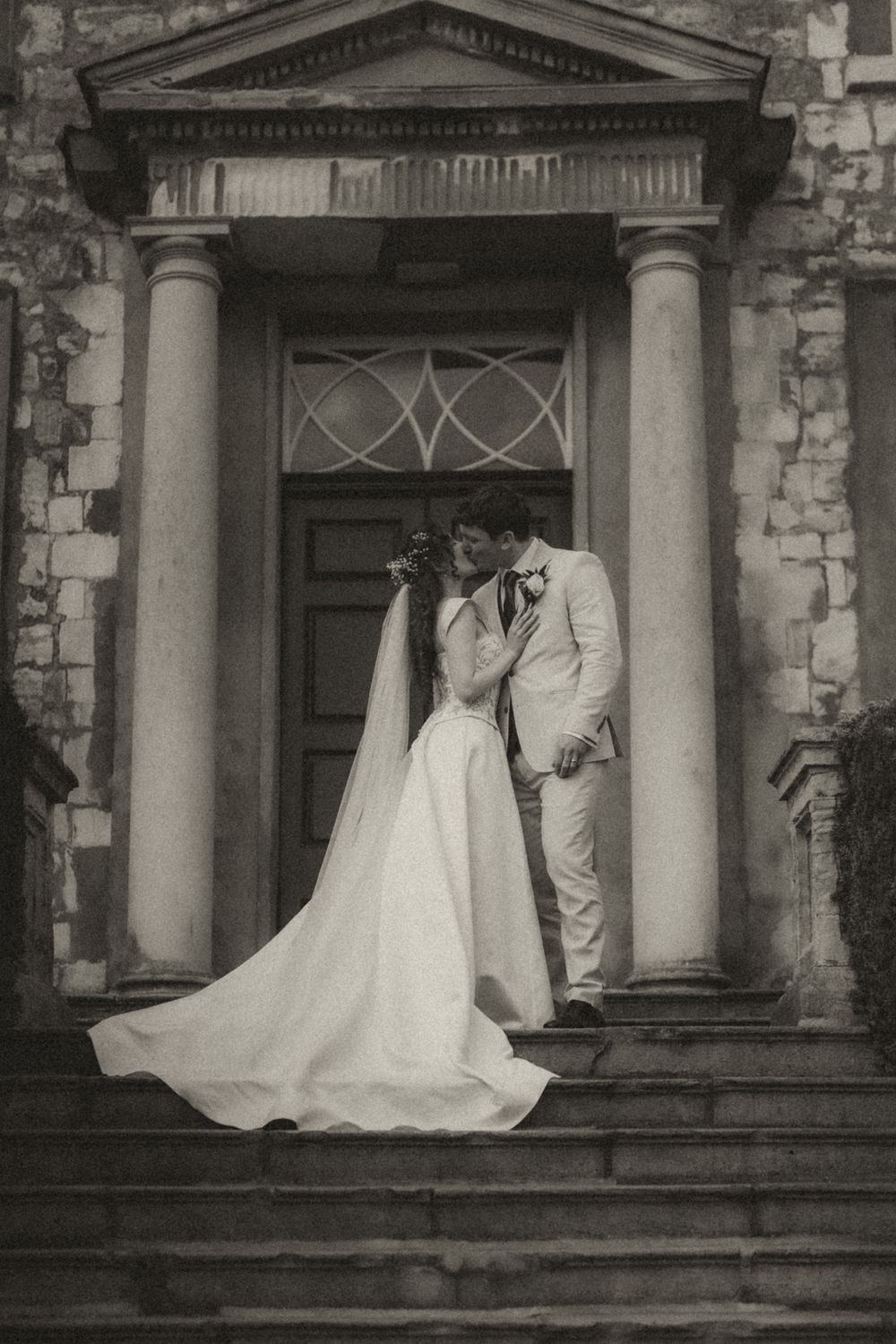 Black and white photo of bride and groom kissing on church steps, wedding dress train flowing.