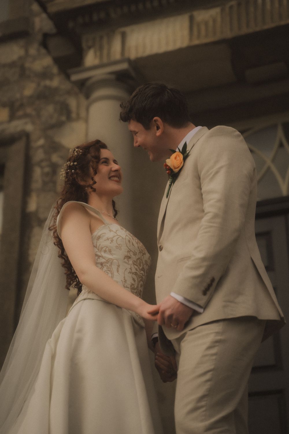 A joyful bride and groom in wedding attire hold hands and share a loving glance outside a stone building.