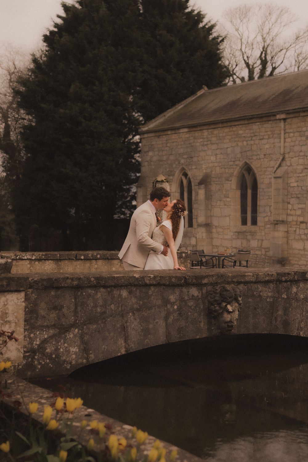A bride and groom share a romantic kiss on a stone bridge beside a historic chapel surrounded by lush trees.