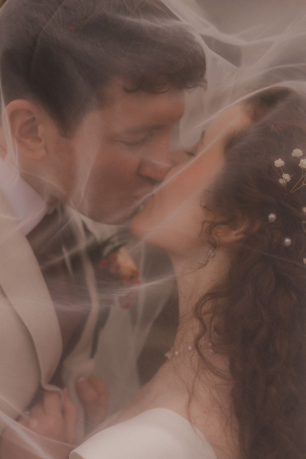 Bride and groom sharing a romantic kiss under a soft bridal veil, with pearl hair accessories and wedding attire.