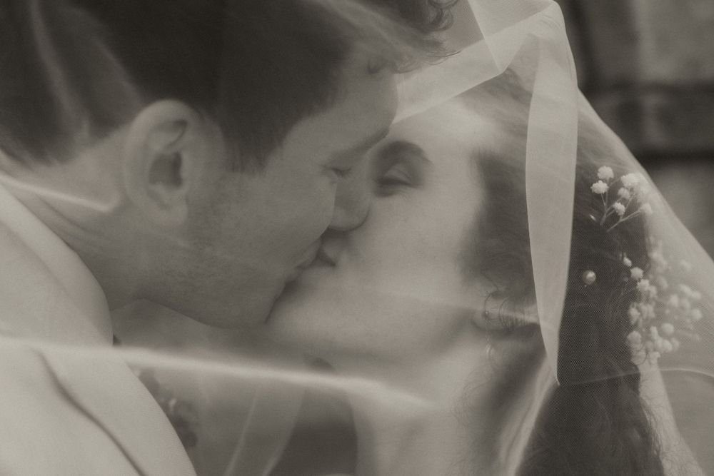 Black and white photo of a bride and groom sharing a romantic kiss beneath a delicate wedding veil.