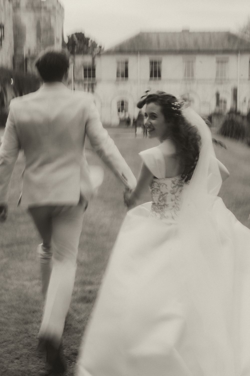 Black and white photo of a bride and groom holding hands, running across a lawn toward a grand estate.