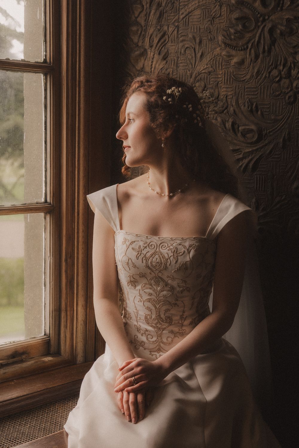 A bride in an embroidered white gown sits by a vintage window, gazing outside with flowers in her curly hair.